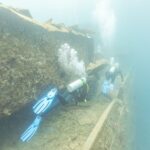Dois mergulhadores passam pelo convés do Thistlegorm ao longo de um corredor, numa água azul leitosa