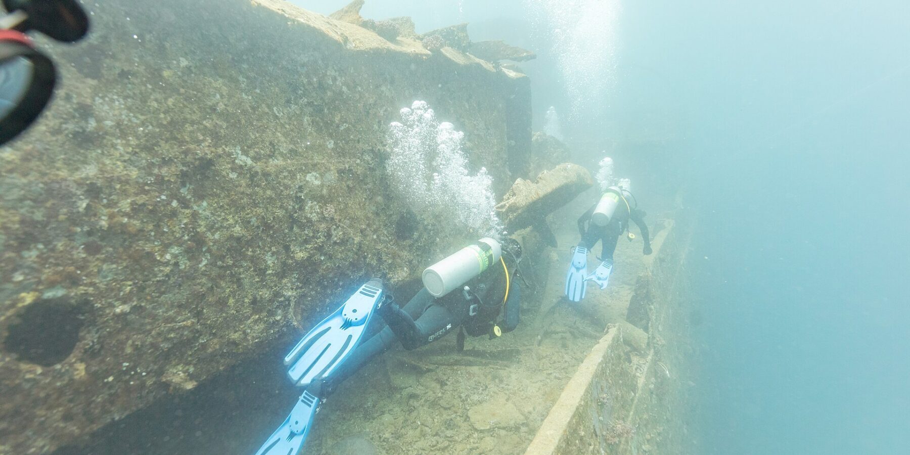 Dois mergulhadores passam pelo convés do Thistlegorm ao longo de um corredor, numa água azul leitosa