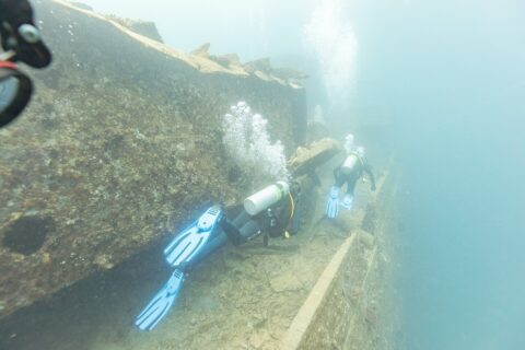 Dois mergulhadores passam pelo convés do Thistlegorm ao longo de um corredor, numa água azul leitosa