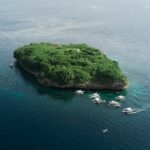 Vista aérea da ilha de Pescador, uma rocha coberta de vegetação verde com seu farol branco, cercada por uma dezena de bancas de mergulho num mar azul-escuro