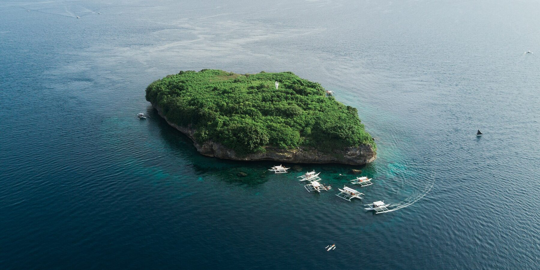 Vista aérea da ilha de Pescador, uma rocha coberta de vegetação verde com seu farol branco, cercada por uma dezena de bancas de mergulho num mar azul-escuro