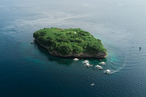 Vista aérea da ilha de Pescador, uma rocha coberta de vegetação verde com seu farol branco, cercada por uma dezena de bancas de mergulho num mar azul-escuro
