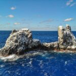Roca Partida vista de drone: uma rocha branqueada pelas aves, partida em dois picos, batida por um mar azul-escuro sob um céu azul com algumas nuvens
