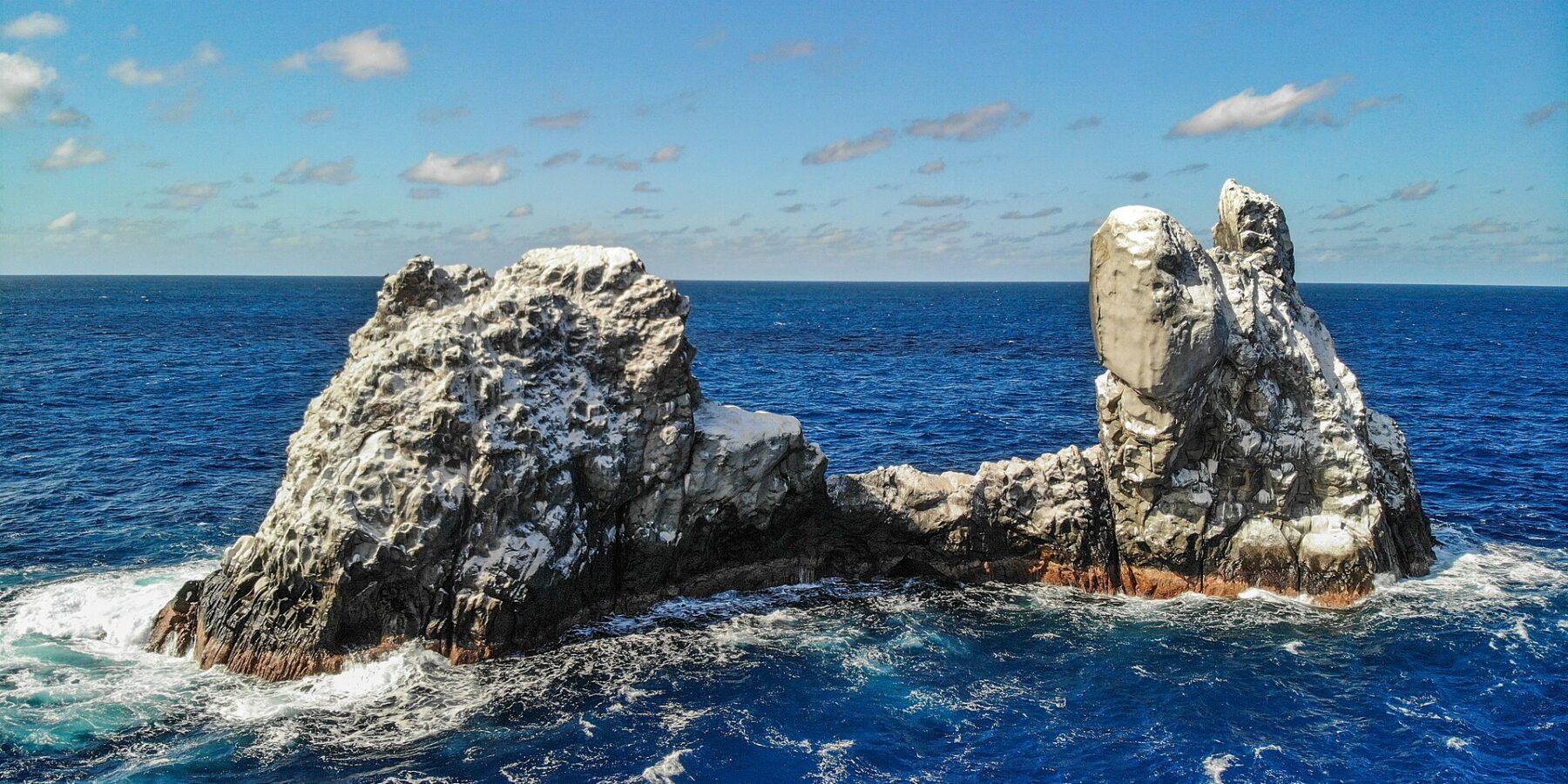 Roca Partida vista de drone: uma rocha branqueada pelas aves, partida em dois picos, batida por um mar azul-escuro sob um céu azul com algumas nuvens