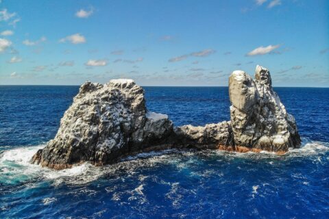 Roca Partida vista de drone: uma rocha branqueada pelas aves, partida em dois picos, batida por um mar azul-escuro sob um céu azul com algumas nuvens