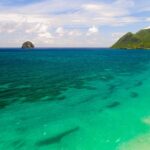 Vista aérea da longa praia de Le Diamant, margeada de água turquesa, com o Rochedo do Diamante ao largo e o morne Larcher, um morro verde, à direita