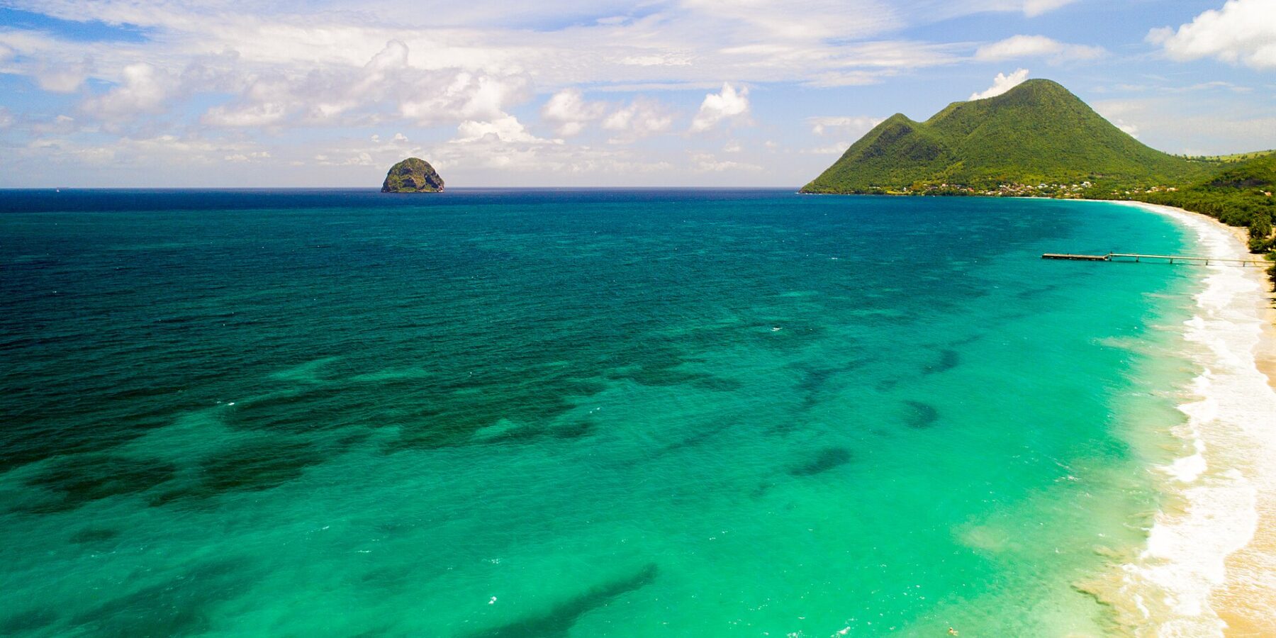 Vista aérea da longa praia de Le Diamant, margeada de água turquesa, com o Rochedo do Diamante ao largo e o morne Larcher, um morro verde, à direita
