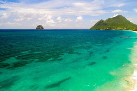 Vista aérea da longa praia de Le Diamant, margeada de água turquesa, com o Rochedo do Diamante ao largo e o morne Larcher, um morro verde, à direita
