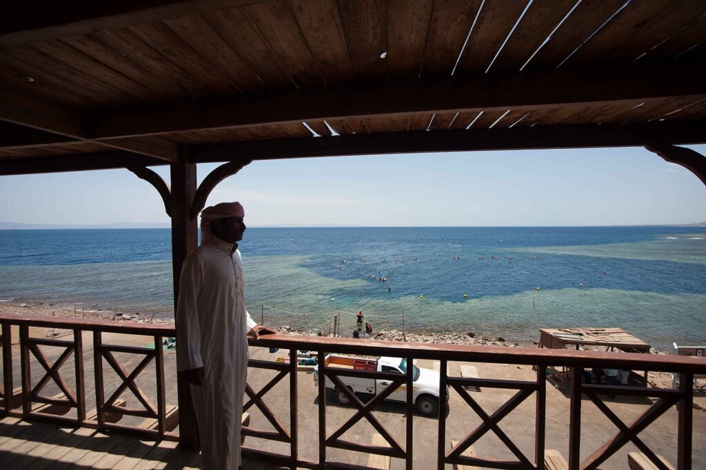 De um deque de madeira, um homem de galabia olha a piscina do Blue Hole, cercada de recife claro, onde nadam banhistas