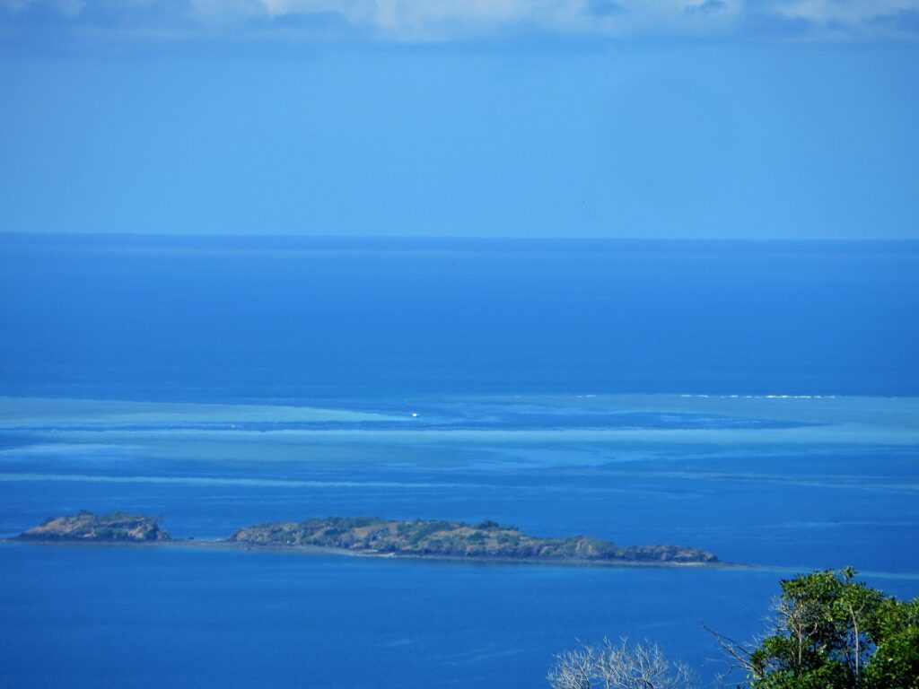 Vista do alto da lagoa azul, duas ilhotas rochosas em primeiro plano e, ao longe, os platôs claros da barreira de coral, onde um barco branco está parado no canal