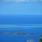 Vista do alto da lagoa azul, duas ilhotas rochosas em primeiro plano e, ao longe, os platôs claros da barreira de coral, onde um barco branco está parado no canal