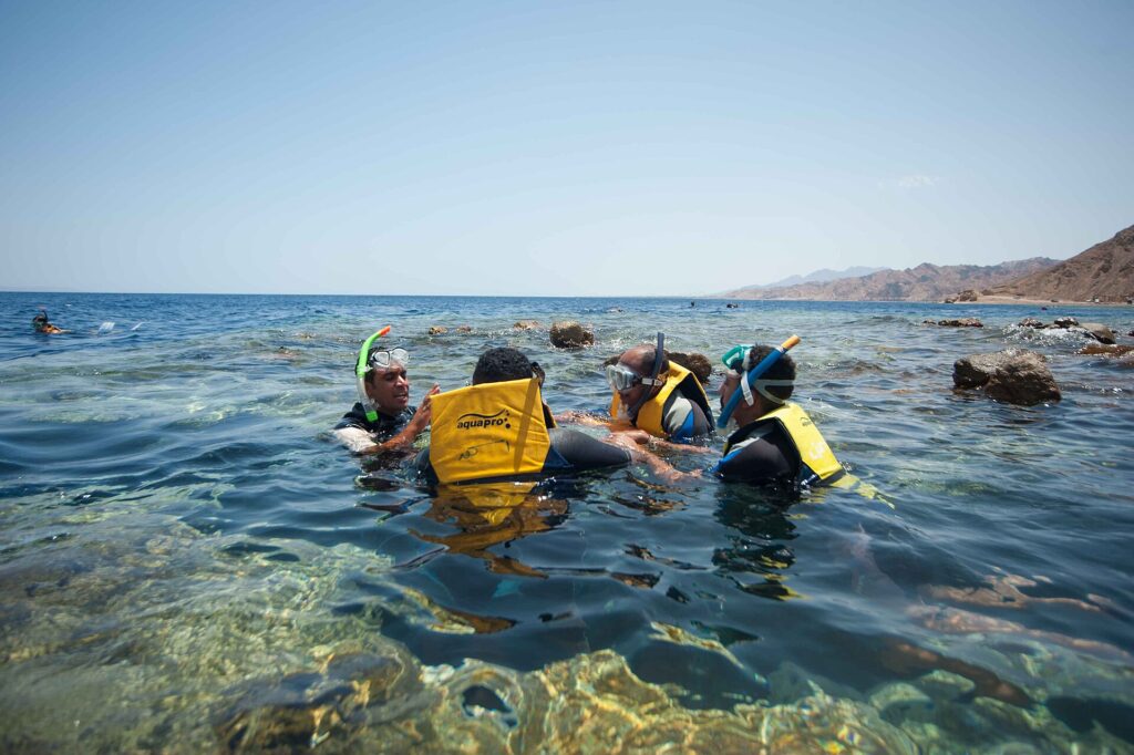 Nadadores de colete amarelo, máscara e snorkel na água clara do Blue Hole, com as montanhas do Sinai atrás