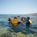 Nadadores de colete amarelo, máscara e snorkel na água clara do Blue Hole, com as montanhas do Sinai atrás